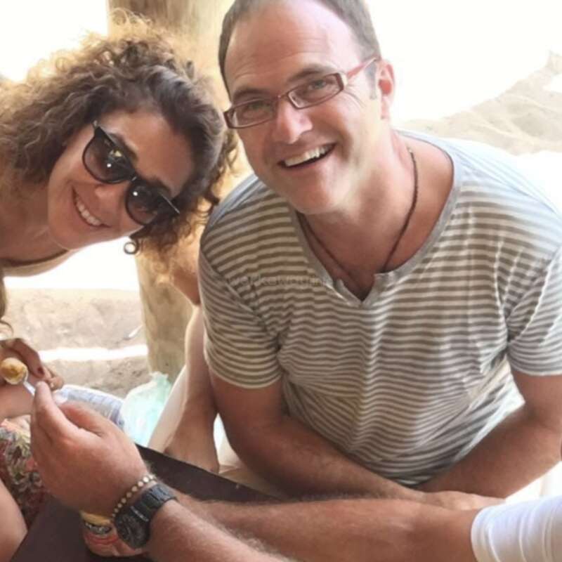 A man and woman smiling brightly at the camera. The man wears glasses and a striped shirt, the woman has curly hair and sunglasses. Casual, cheerful moment shared.