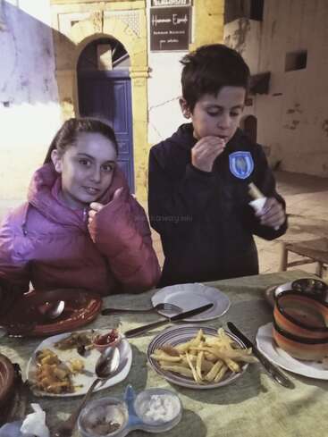 Two children are sitting at a table outdoors, enjoying a meal with fries, vegetables, and dips. The girl smiles with a thumbs up, looking happy.