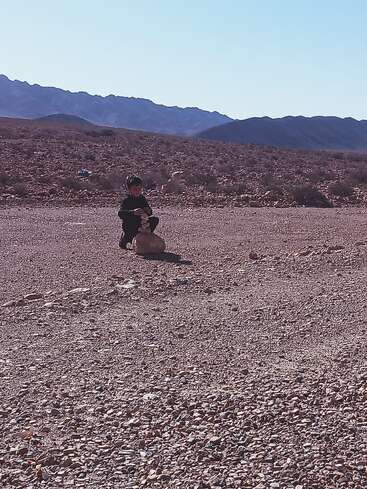 A child sits alone in a rocky, desert landscape, stacking stones on top of a large rock. Mountains are visible in the background under a clear sky.