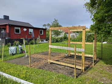 La imagen muestra una pequeña parcela de jardín vallada con postes de madera en un patio trasero cubierto de hierba. Al fondo se ve una casa roja, herramientas de jardinería y árboles.