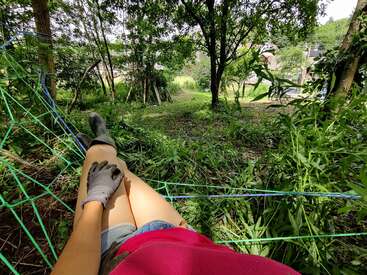 Une personne se détend sur un hamac en filet dans un jardin verdoyant. Elle porte des gants, un short et une chemise rose, et profite d'un moment paisible en plein air.