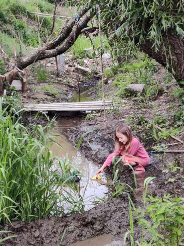 Une jeune fille en tenue rose joue près d'un ruisseau boueux, faisant flotter des canards en peluche. De la verdure luxuriante et un pont en bois rustique rehaussent la scène naturelle.