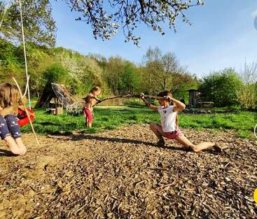 Trois enfants jouent en plein air avec des bâtons, faisant semblant de se battre à l'épée. La lumière du soleil, l'herbe verte, les arbres et une structure en bois créent une atmosphère naturelle, joyeuse et ludique au printemps.