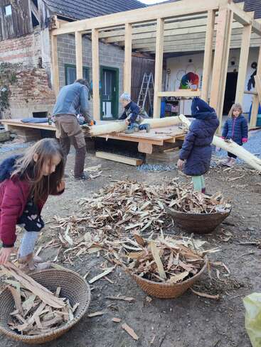 Quatre enfants et un homme travaillent ensemble à l'extérieur, rassemblant des lamelles de bois dans des paniers à côté d'un bâtiment en bois partiellement construit, suggérant un projet communautaire de construction ou de restauration.