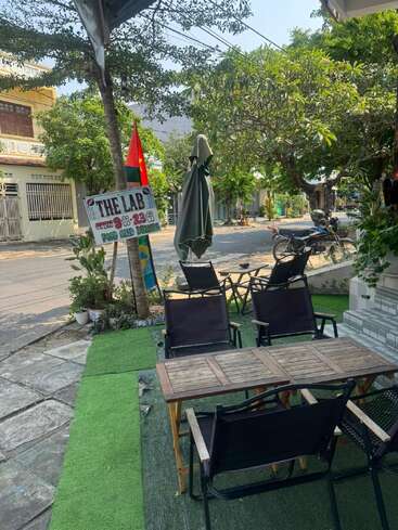 Outdoor seating area with wooden tables, black chairs, and closed umbrella. Green artificial grass flooring, "The Lab" sign, plants, nearby street, trees, and parked motorbike.