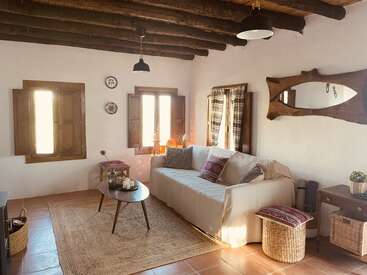 This cozy rustic living room features wooden ceiling beams, warm sunlight, a comfortable sofa with pillows, woven baskets, a unique mirror, and earthy decorative elements.