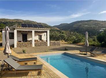 A small white villa with solar panels sits by a swimming pool, surrounded by lounge chairs and umbrellas, overlooking green hills and a clear blue sky.