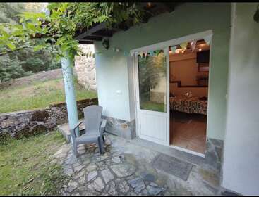 Una acogedora entrada de casa de campo con puerta de cristal, patio de piedra, silla de plástico, vegetación exterior, luces de cuerda y banderines. El interior revela una cama y decoración rústica.