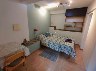 Un acogedor dormitorio con una cama con ropa de cama de flores, almohadas, una toalla doblada, un pequeño escritorio con una silla, estanterías, una lámpara de pared y suelo de baldosas.
