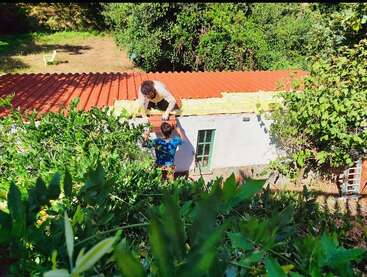 Dos personas trabajan en el tejado de un pequeño edificio, rodeadas de una exuberante vegetación. Una persona está de pie abajo, la otra arrodillada arriba, colaborando.