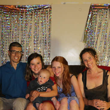 A imagem mostra uma família de cinco pessoas posando para uma foto, com dois adultos e três crianças, em um cenário de cortinas prateadas e uma parede branca.