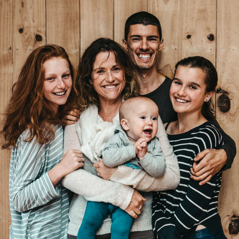 A imagem mostra uma família de cinco pessoas posando para uma foto em frente a uma parede de madeira, com a mãe segurando um bebê e três crianças ao seu lado.