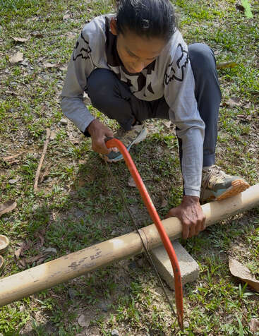 A person kneels on the grass, using a handsaw to cut a bamboo pole supported by a concrete block, wearing casual clothes and focused on the task.