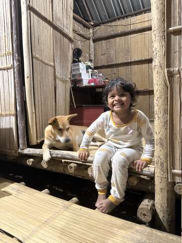 A cheerful child in pajamas sits on a wooden porch next to a brown dog inside a rustic bamboo house, both appearing comfortable and happy together.