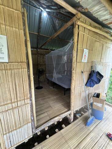 This image shows a rustic bamboo hut with a metal roof, basic bedding, mosquito net, broom, cardboard box, fan, and various items hanging by the doorway.