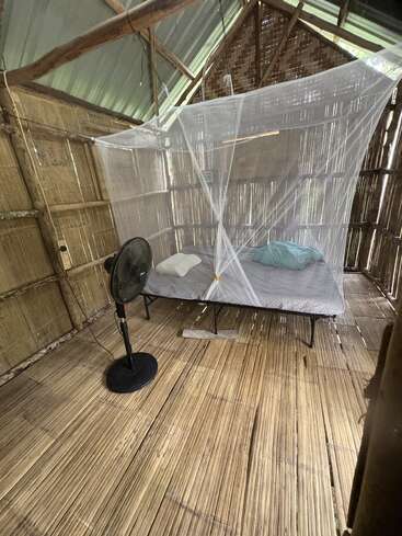 This image shows a simple bamboo room with a single bed covered by a mosquito net, a standing electric fan, wooden slatted floor, and rustic walls.
