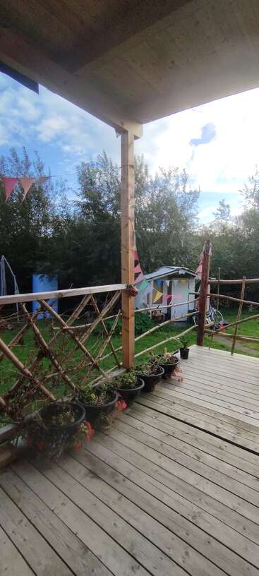Un porche en bois avec des plantes en pot et des balustrades rustiques, décoré de guirlandes colorées sous un ciel partiellement nuageux, surplombe l'herbe verte et les arbres en arrière-plan.