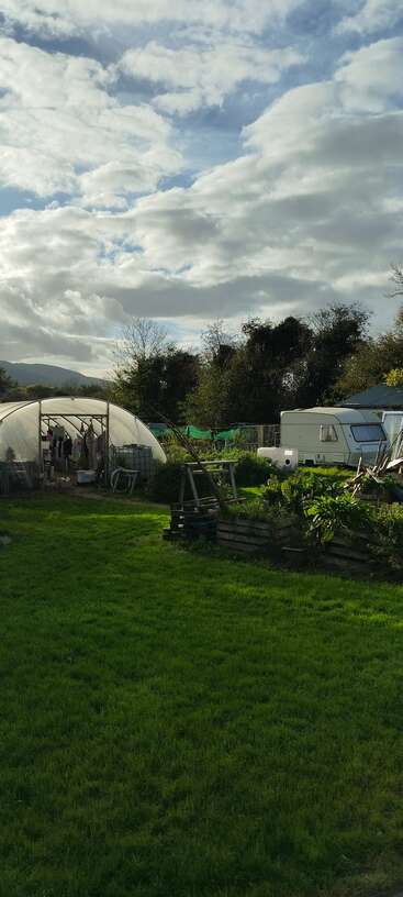 Un jardin verdoyant avec des lits surélevés, un polytunnel et un séchoir à linge. Une caravane blanche se trouve à proximité, le tout sous un ciel partiellement nuageux et ensoleillé.