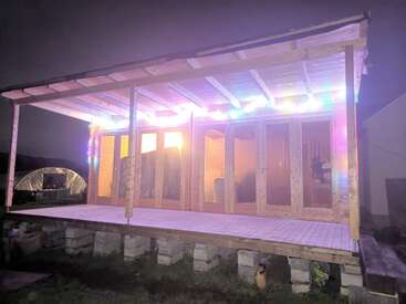 L'image représente une structure en bois avec une terrasse et un toit, avec une porte et des fenêtres, située dans une cour la nuit, avec une petite tente à l'arrière-plan.