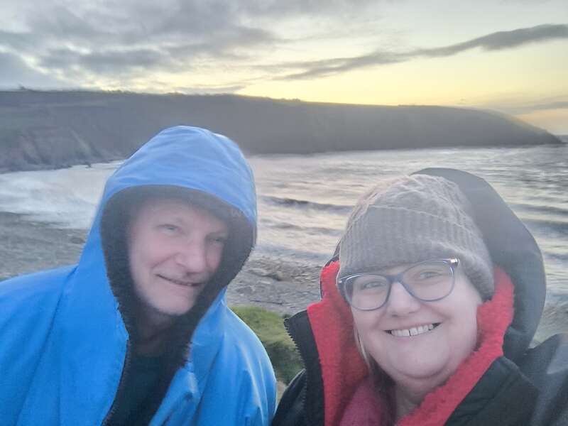 L'image montre deux personnes en vêtements d'hiver debout sur une plage rocheuse, avec une falaise et un plan d'eau derrière eux, sous un ciel nuageux.