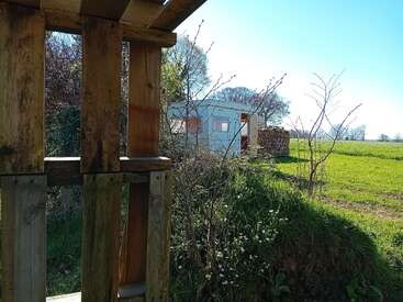 A wooden pallet frames a scenic rural field, with a white shed and stacked firewood in the distance, under a clear blue sky and sunlight.