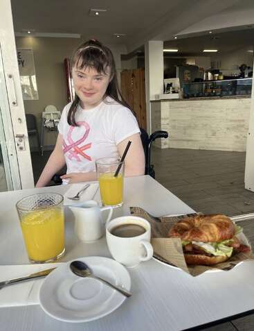 Eine junge Frau sitzt an einem Cafétisch mit Orangensaft, Kaffee und einem Croissant-Sandwich und lächelt herzlich. Die Atmosphäre im Innenbereich ist gemütlich und einladend.