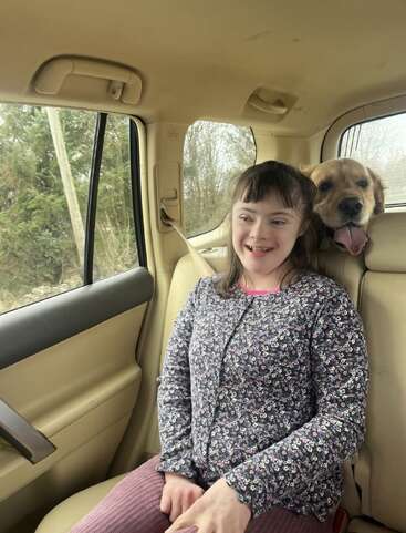 Ein lächelndes Mädchen sitzt auf dem Rücksitz eines Autos und trägt ein geblümtes Oberteil. Neben ihr streckt ein fröhlicher Hund den Kopf nach vorne und lässt die Zunge heraushängen.