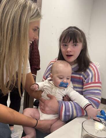 Eine junge Frau lächelt ein Mädchen an, das ein Baby mit einem blauen Schnuller im Arm hält. Das Baby streckt die Hände aus, und die Gruppe erlebt gemeinsam einen schönen, fröhlichen Moment.