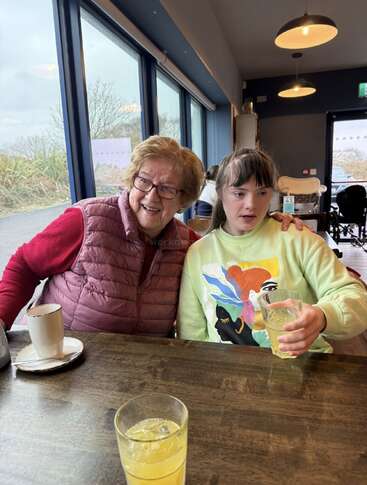 Eine ältere Frau und ein junges Mädchen sitzen zusammen an einem Cafétisch, lächeln und genießen ihre Getränke. Durch die großen Fenster fällt Tageslicht herein. Gemütliche Atmosphäre.