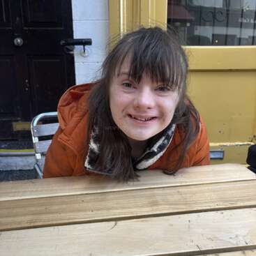A young girl with long brown hair, wearing an orange jacket, smiles warmly while sitting at a wooden table outside a café with yellow and black doors.