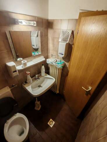 Este pequeño cuarto de baño cuenta con un inodoro, un lavabo, un espejo, un dispensador de jabón, productos de limpieza, papel higiénico, un dispensador de toallas de papel, un radiador, paredes alicatadas y una puerta de madera.