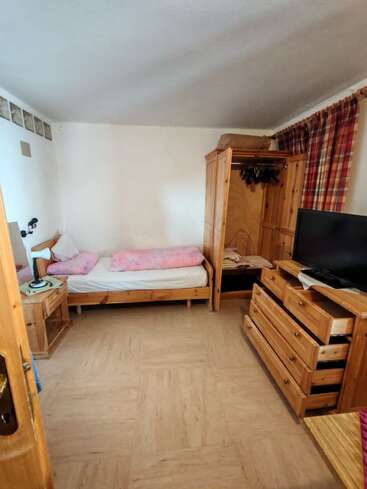 Esta acogedora habitación cuenta con muebles de madera, una cama individual con ropa de cama rosa, un armario abierto, una cómoda, un televisor, cortinas a cuadros y un suelo de colores neutros.