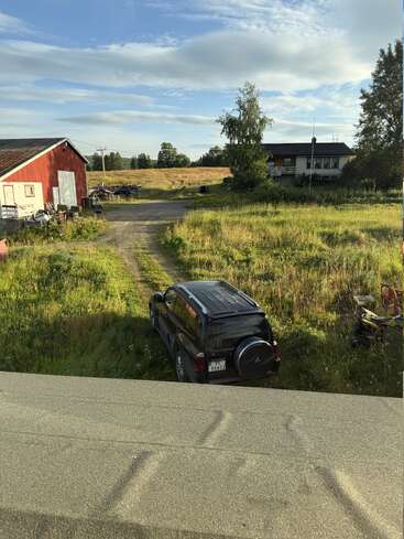 A black SUV is parked on grassy land near a rural house and red barn. The sky is blue with scattered clouds, and fields surround the scene.