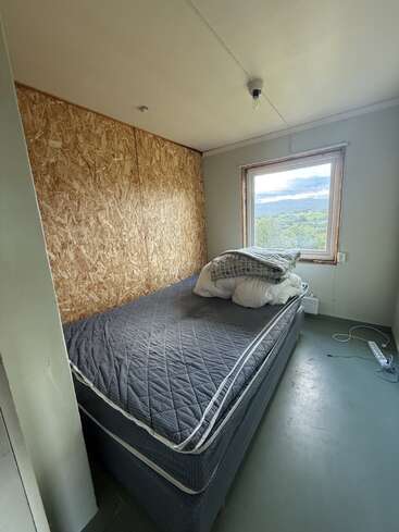 A small, minimalist bedroom with a bare mattress, pillows, and blanket. Plywood accent wall, single lightbulb, window with scenic view, and power strip on the floor.