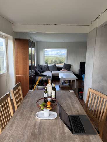 This image shows a cozy, well-lit dining and living area. A wooden table with a laptop, fruit, and condiments faces a comfortable gray sectional sofa.