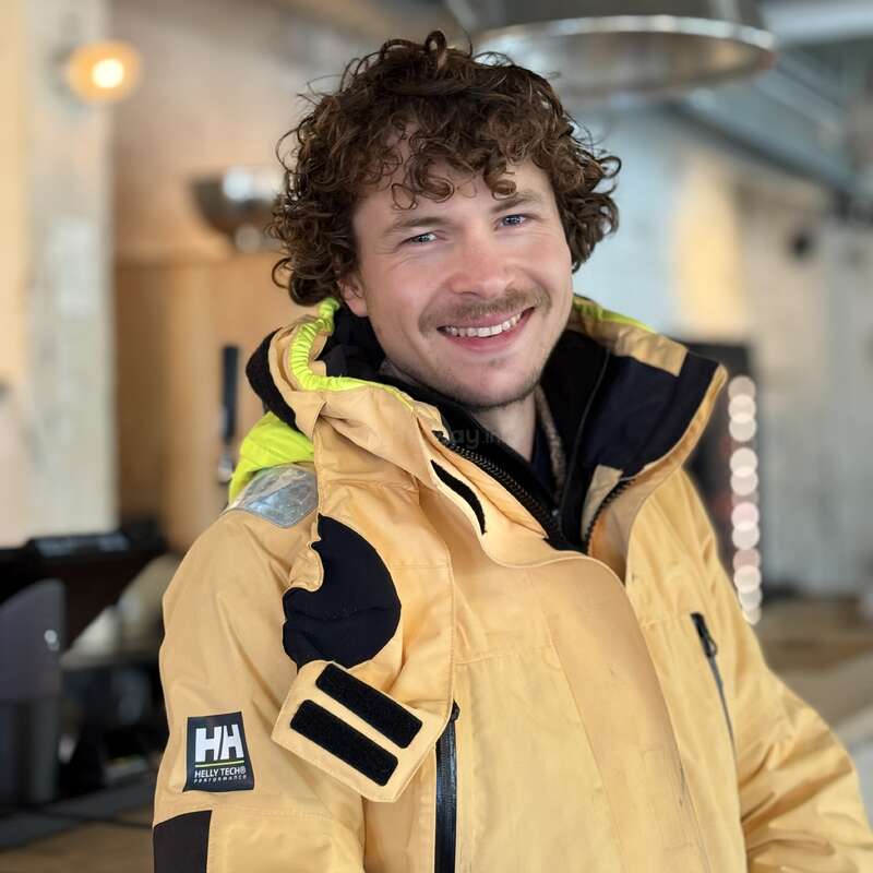 A smiling man with curly hair wears a yellow Helly Hansen jacket indoors. The background is softly blurred, featuring warm lighting and industrial-style decor elements.