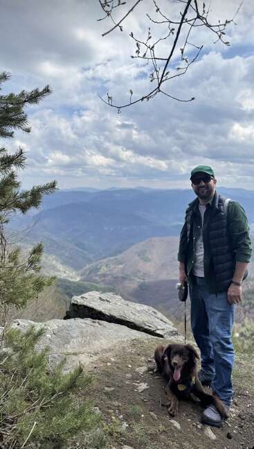 A man wearing sunglasses and a green cap stands on a mountain trail with his happy dog. Scenic mountains, cloudy sky, and pine trees surround them.