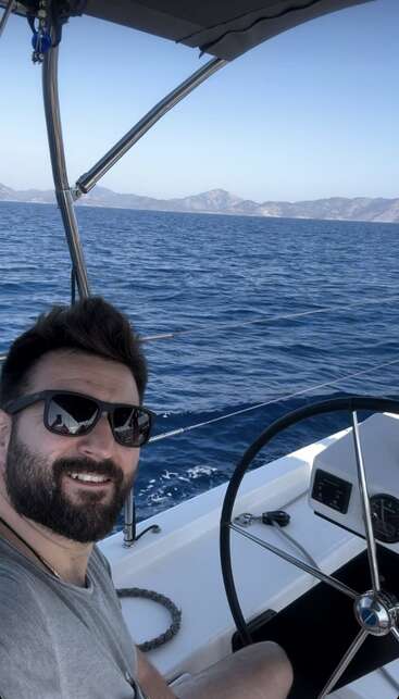 A bearded man in sunglasses smiles while steering a boat on calm blue sea, with mountains and clear sky in the background, enjoying a sunny day.