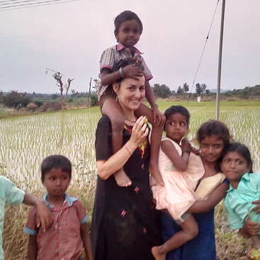 The image depicts a woman in a black dress carrying a child on her shoulders, surrounded by children in a field with trees and a cloudy sky in the background.