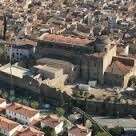 L'image représente une vue aérienne d'une ville, montrant un grand bâtiment en pierre avec un toit en tuiles rouges, entouré de bâtiments plus petits et d'arbres.