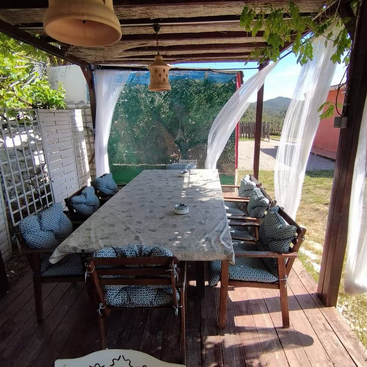 Ein gemütlicher Essbereich im Freien mit einem Holztisch, gepolsterten Stühlen, weißen Vorhängen, Hängelampen und einer grünen Kulisse unter einem Pergoladach.