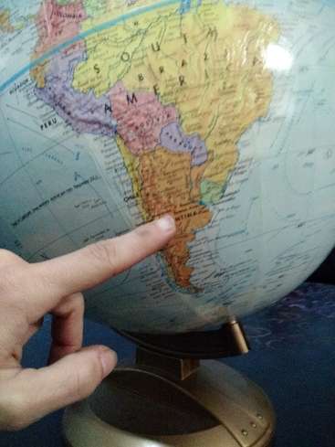 Una mano señala América del Sur en un globo terráqueo, con los países del continente visibles sobre un fondo azul. El globo está sobre un soporte de bronce.