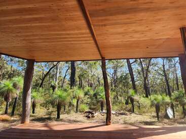 Una cubierta de madera con un gran tejado da a un apacible bosque lleno de árboles altos y frondosos céspedes verdes bajo la brillante luz del sol de la tarde.