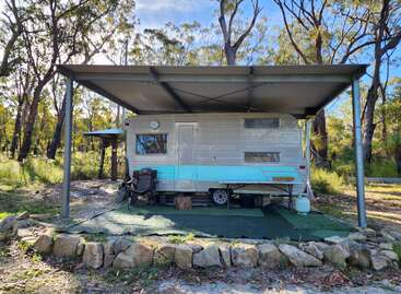 Una caravana de época con una franja turquesa descansa bajo un refugio metálico en un apacible entorno forestal. En el exterior descansan sillas de camping y diversos objetos.