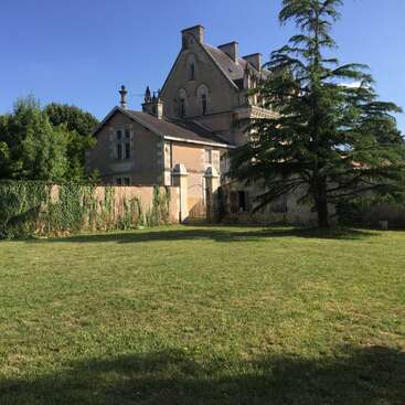 The image depicts a large, tan-colored house with a brown roof, situated behind a grassy field and a stone wall, surrounded by trees and a clear blue sky.