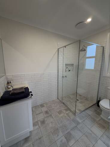 This image shows a modern bathroom with white subway tiles, a glass shower enclosure, a small vanity with towels, tiled floor, toilet, and natural light.