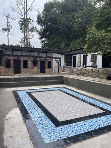 Esta imagem mostra uma piscina de azulejos vazia em frente a edifícios rústicos de tijolos, cercada por árvores verdes exuberantes e um muro de pedra, sob um céu nublado.