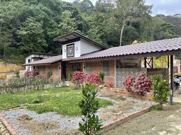 Esta imagem mostra uma casa rústica de tijolos com telhado de telha, cercada por uma vegetação exuberante, plantas floridas e árvores, aninhada na borda de uma floresta.