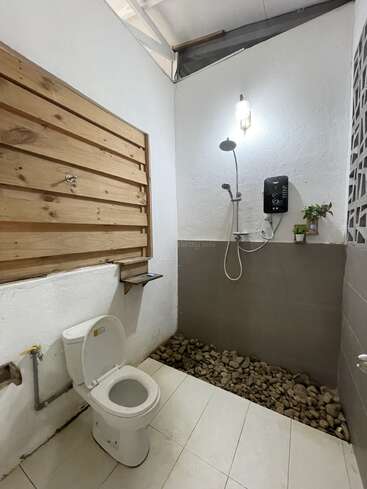 Esta imagem mostra um banheiro minimalista com vaso sanitário branco, chuveiro aberto, piso de cascalho, detalhes em madeira na parede, plantas e teto parcialmente aberto para luz natural.