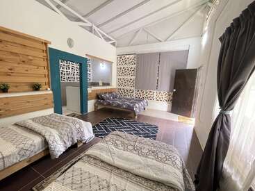 Um quarto moderno e aconchegante com três camas, roupa de cama estampada, painéis decorativos de madeira, tapete geométrico, cortinas, plantas de interior, janelas grandes e iluminação aconchegante, criando uma atmosfera acolhedora.